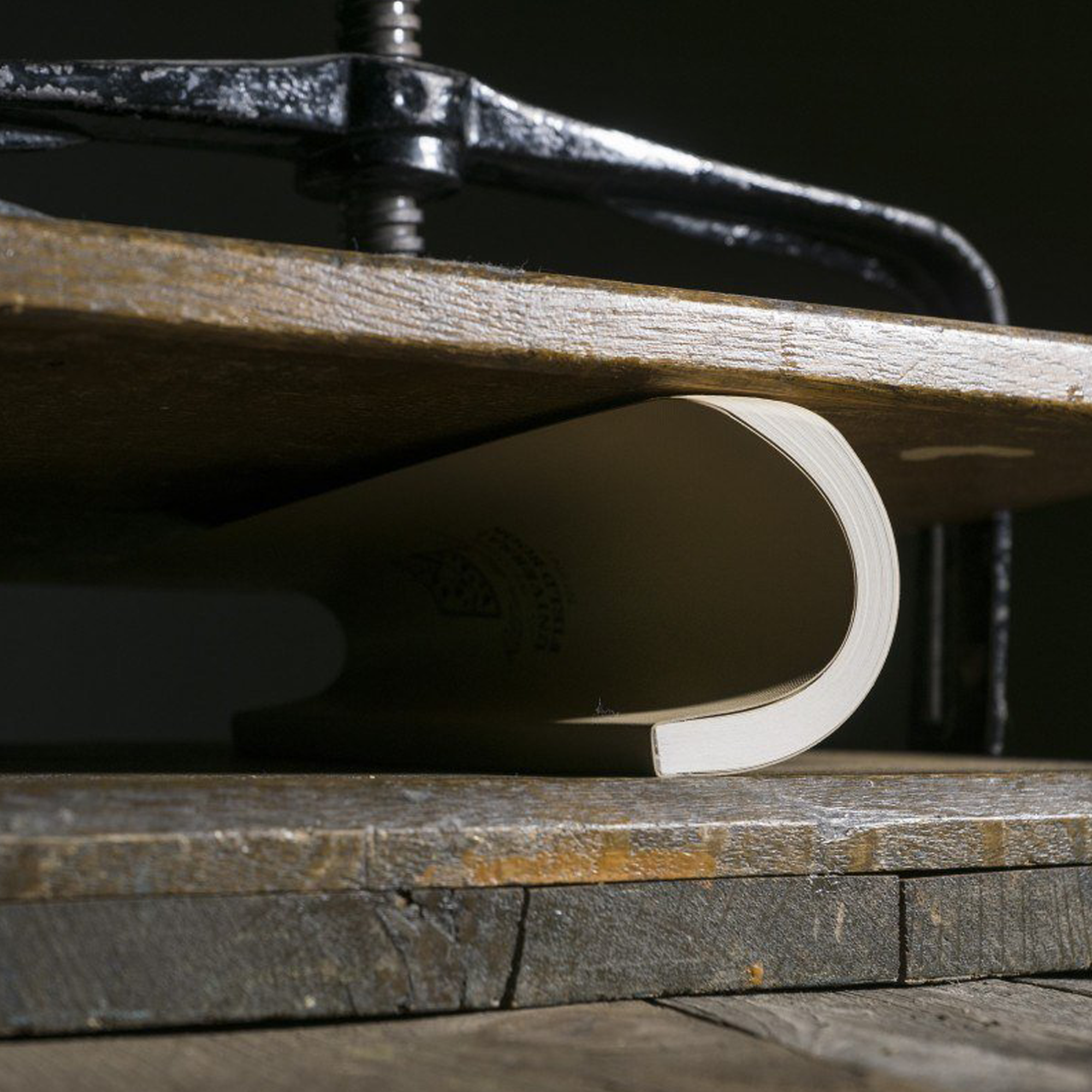 A close-up of a curved book under a black metal clamp on a rustic wooden surface.