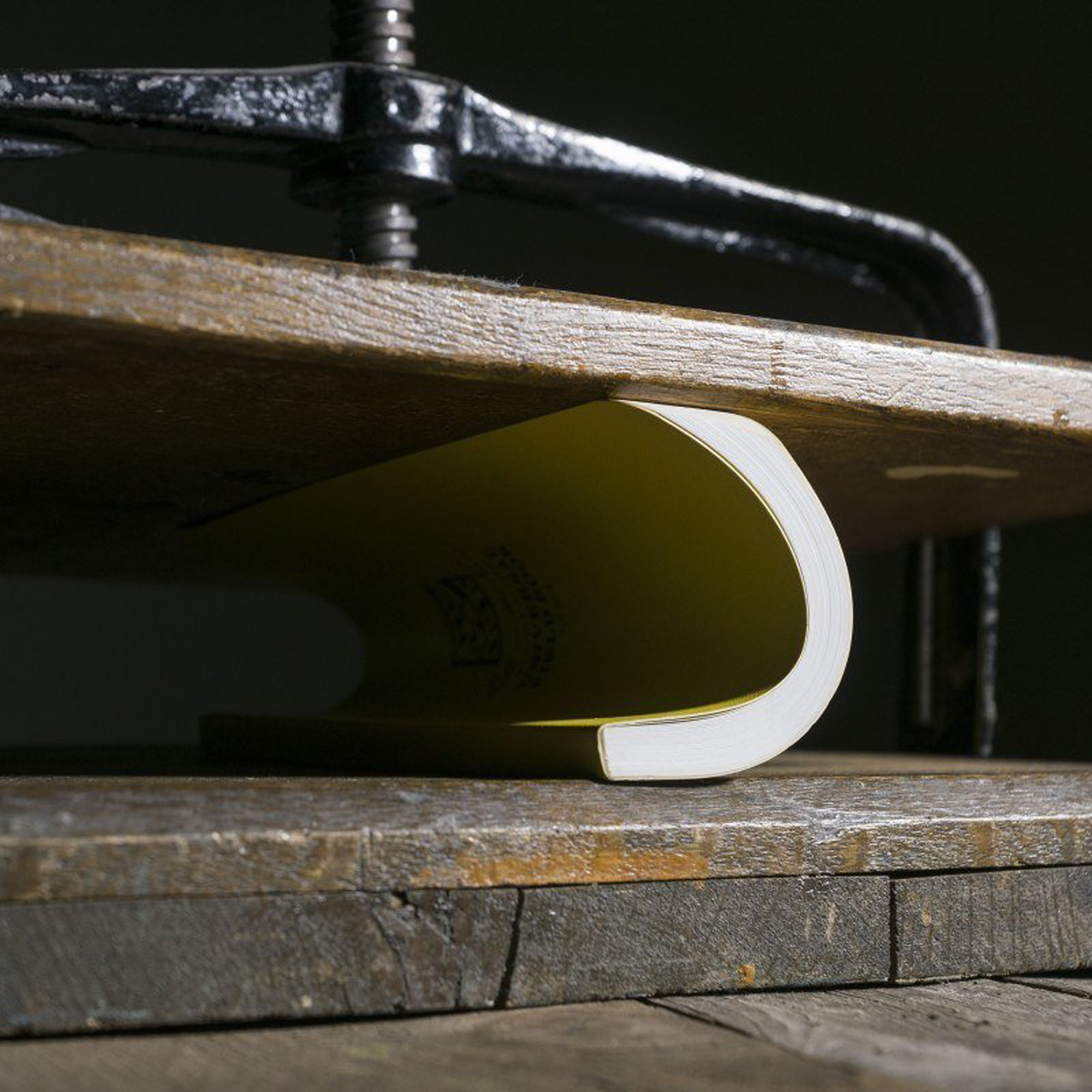 A close-up of a curved book under a black metal clamp on a rustic wooden surface.