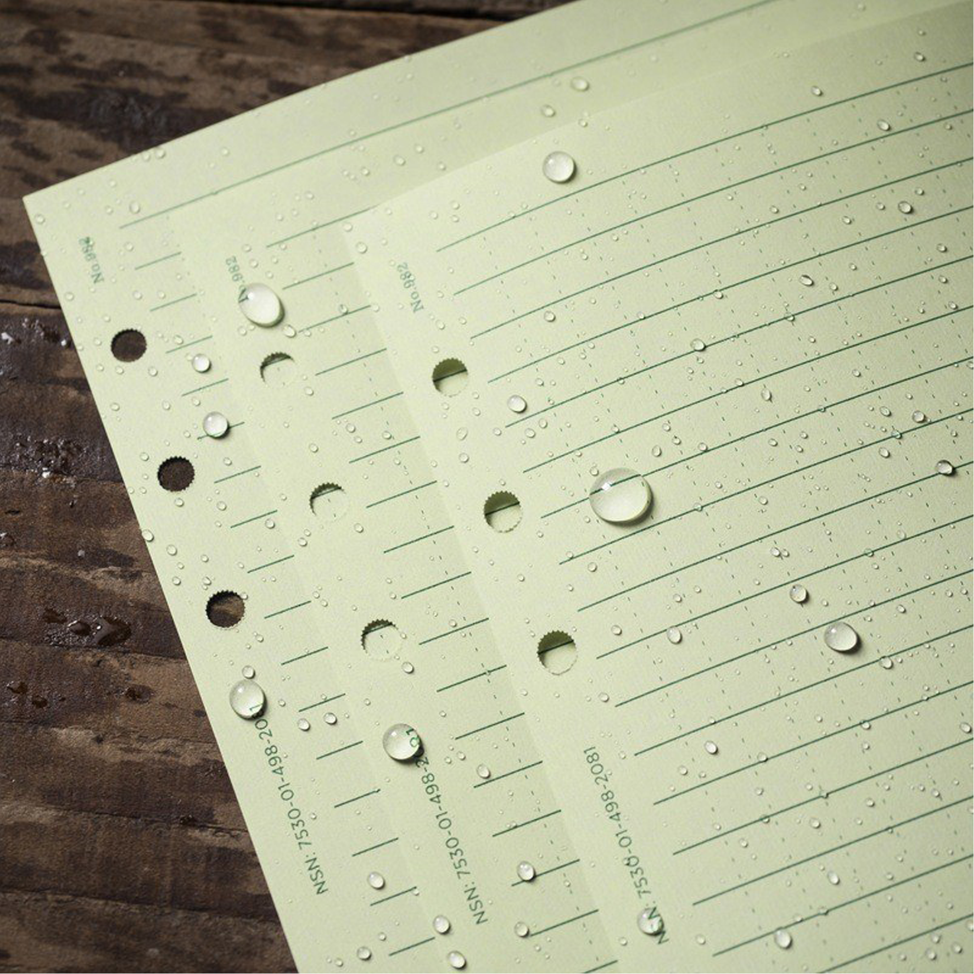 Green lined paper sheets with water droplets on a dark wooden surface, showing water resistance.