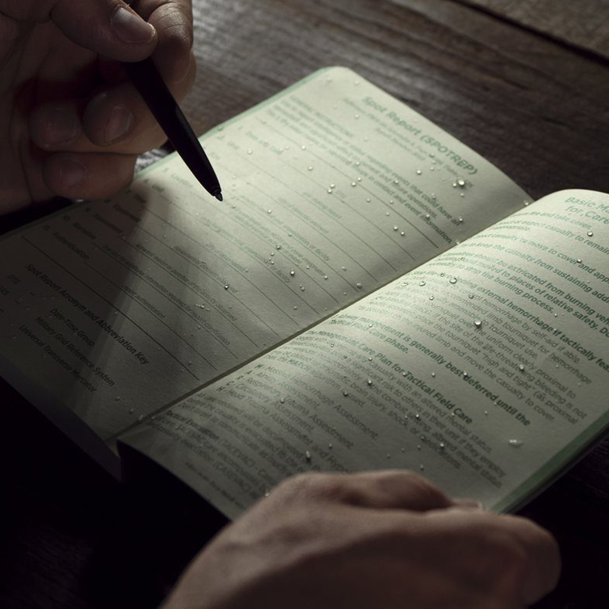 An open notebook lying on a dark wooden table, with water droplets on the pages. A hand holds a black pen